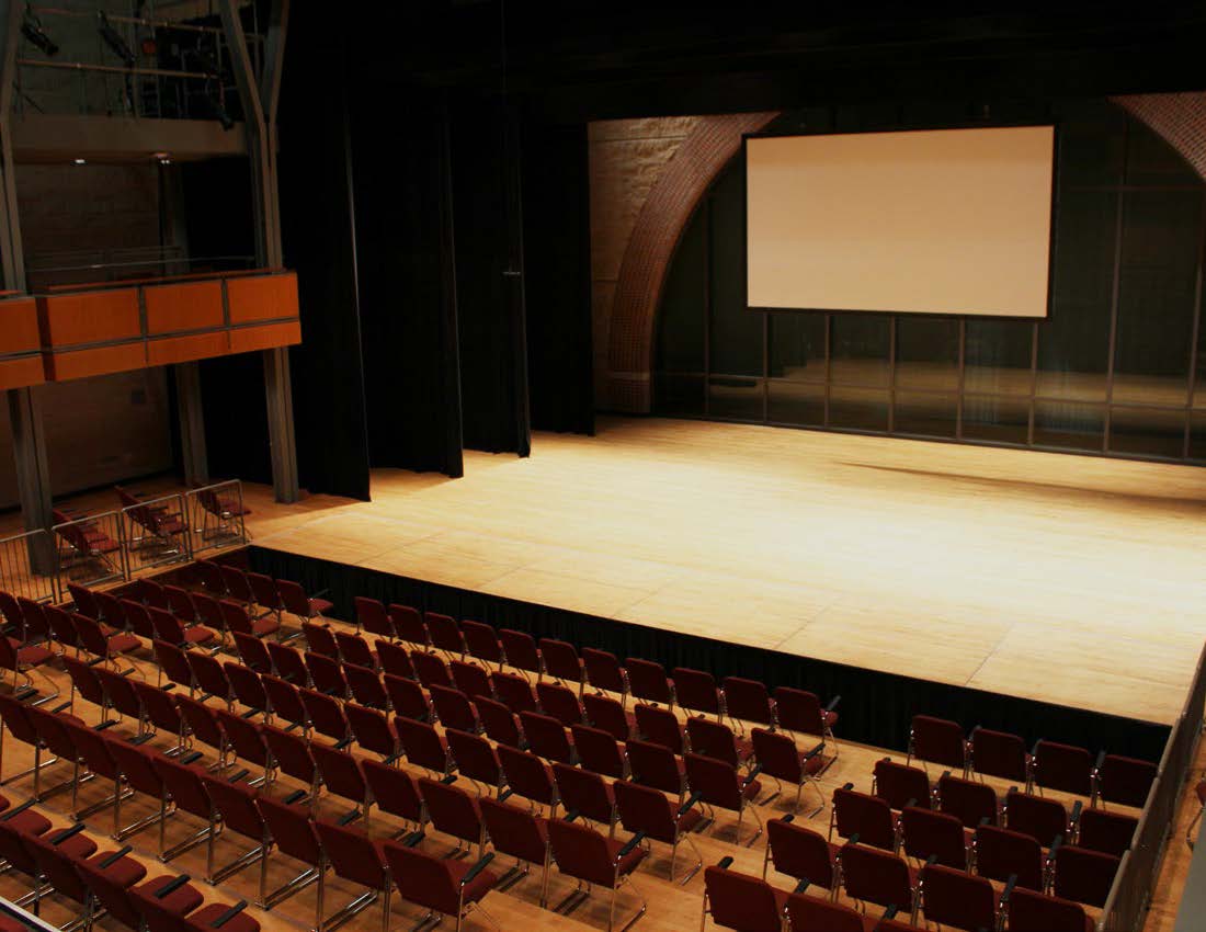 Empty auditorium with rows of red seats facing a wooden stage and a large projection screen.